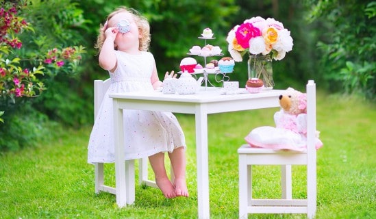 Adorable funny toddler girl with curly hair wearing a colorful dress on her birthday playing tea party with a teddy bear doll, toy dishes, cup cakes and muffins in a sunny summer garden