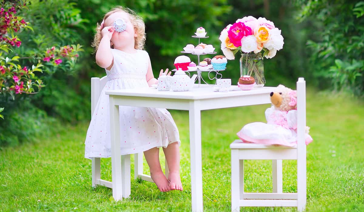 Adorable funny toddler girl with curly hair wearing a colorful dress on her birthday playing tea party with a teddy bear doll, toy dishes, cup cakes and muffins in a sunny summer garden